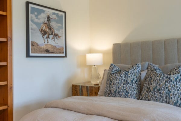 Close up of bed with decorative pillows, side table with lamp, artwork of a bucking horse with rider hangs on the wall, and built-in wall shelves.